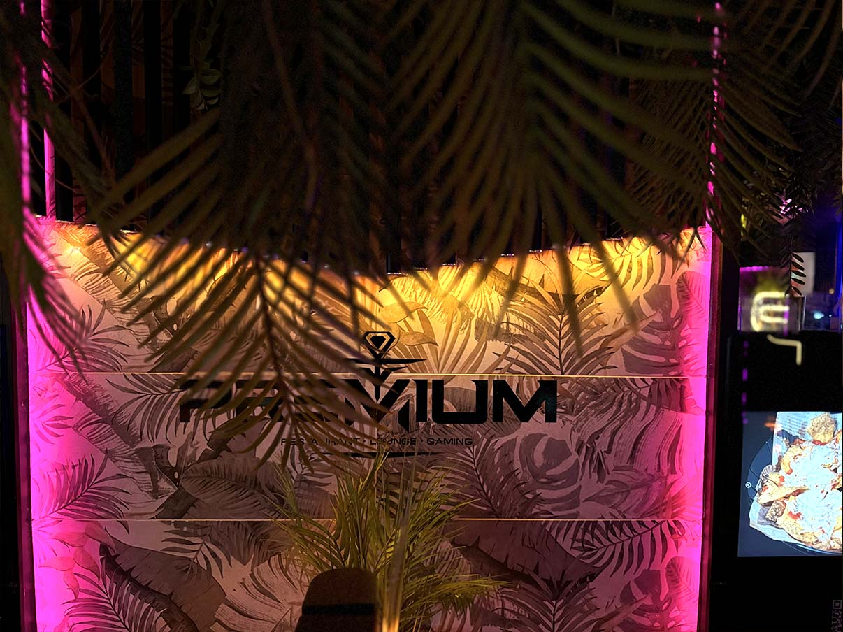 Vor einer mit floralen Mustern und bunten LED-Lichtern beleuchteten Wand in der hookah hafencity hängen palmähnliche Blätter herab. Das Wort "Premium" ist prominent auf der dekorierten Wand zu sehen.