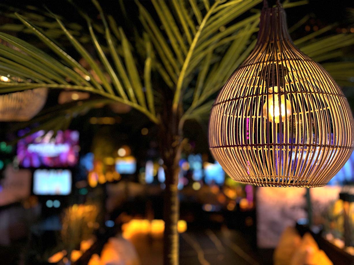 Eine stimmungsvolle Szene in einer shisha bar hafencity, bei der ein kunstvoller, runder Lampenschirm aus Holzgeflecht im Vordergrund warmes Licht abgibt. Im Hintergrund sind Palmenblätter, bunte Lichter und verschwommene Bildschirme zu erkennen.