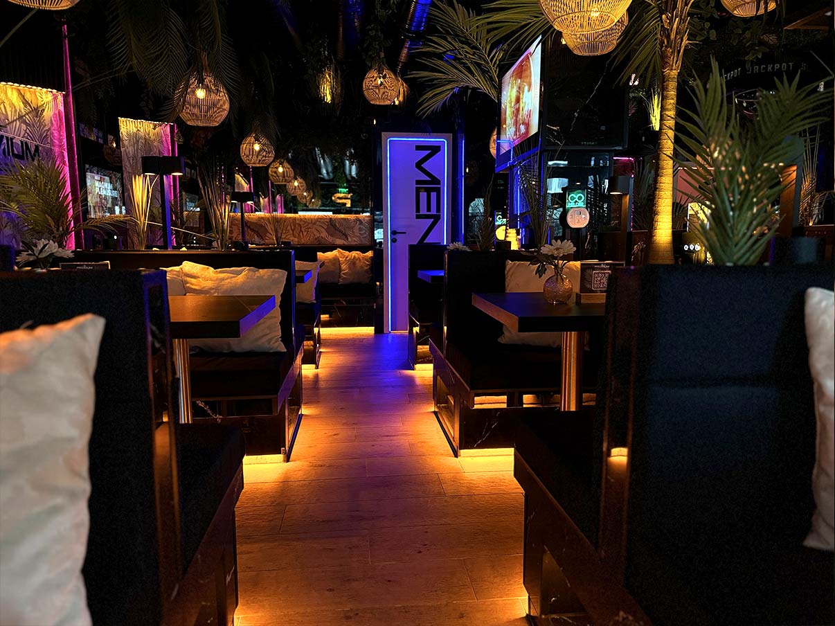 Ein Blick ins Innere der shisha bar hamburg hafencity: Mehrere Sitzecken mit dunklen Polstern, leuchtenden Bodenakzenten, dekorativen Lampenschirmen und tropischen Pflanzen vermitteln ein modernes, gemütliches Ambiente.