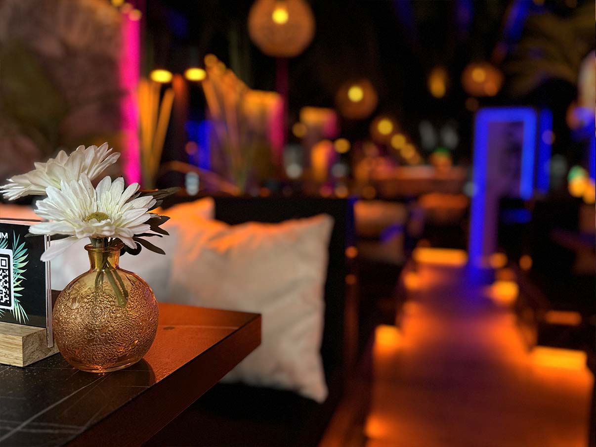 In einer stilvollen shisha lounge hafencity steht im Vordergrund eine kleine goldfarbene Vase mit weißen Blüten auf einem Tisch, während im Hintergrund weiche Kissen und warmes Licht ein gemütliches Ambiente andeuten.