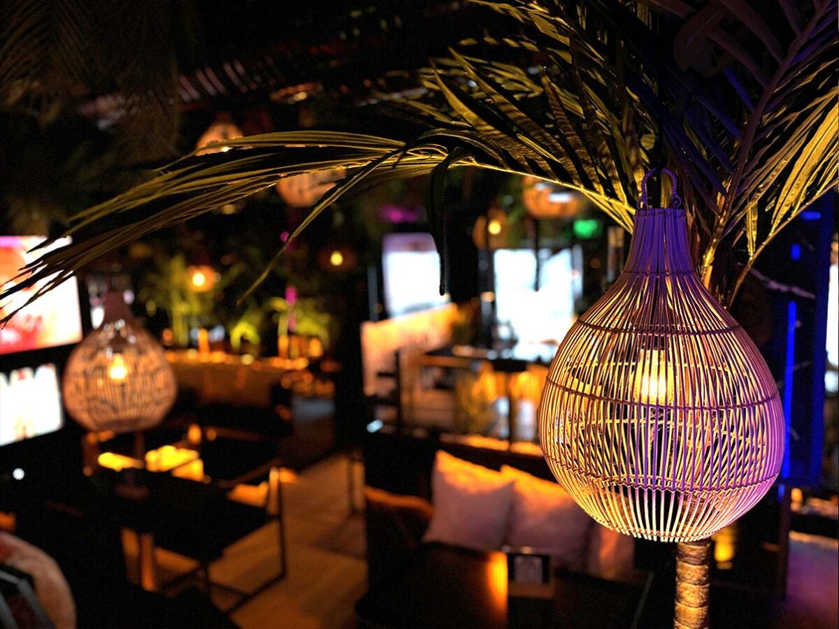 Ein runder, geflochtener Lampenschirm hängt in einer stimmungsvoll beleuchteten shisha plae hafencity, umgeben von palmähnlichen Blättern und warmen Lichtakzenten.