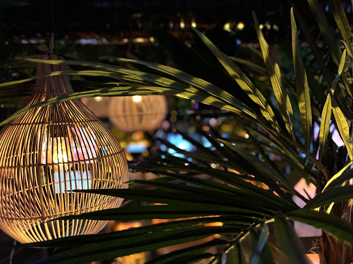 Ein dekorativer, geflochtener Lampenschirm hängt vor üppigen Palmenblättern, während warme Lichteffekte und bunte Farbakzente im Hintergrund die stimmungsvolle shisha bar hafencity andeuten.